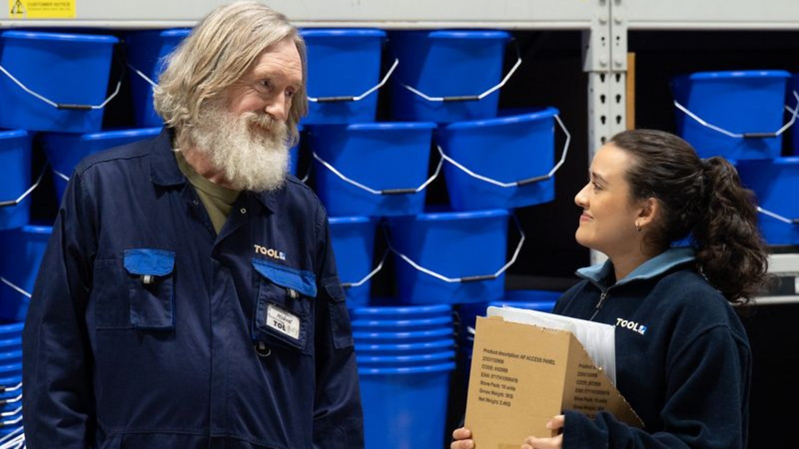 A man and a young woman in navy shop uniforms stand in front of a stack of blue mop buckets. She holds a cardboard box.