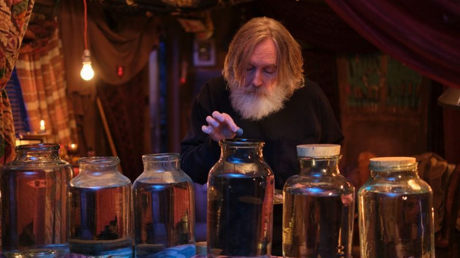 A bearded man holds something above a glass jar in a darkened room — the homunculi shed scene from Small Prophets.